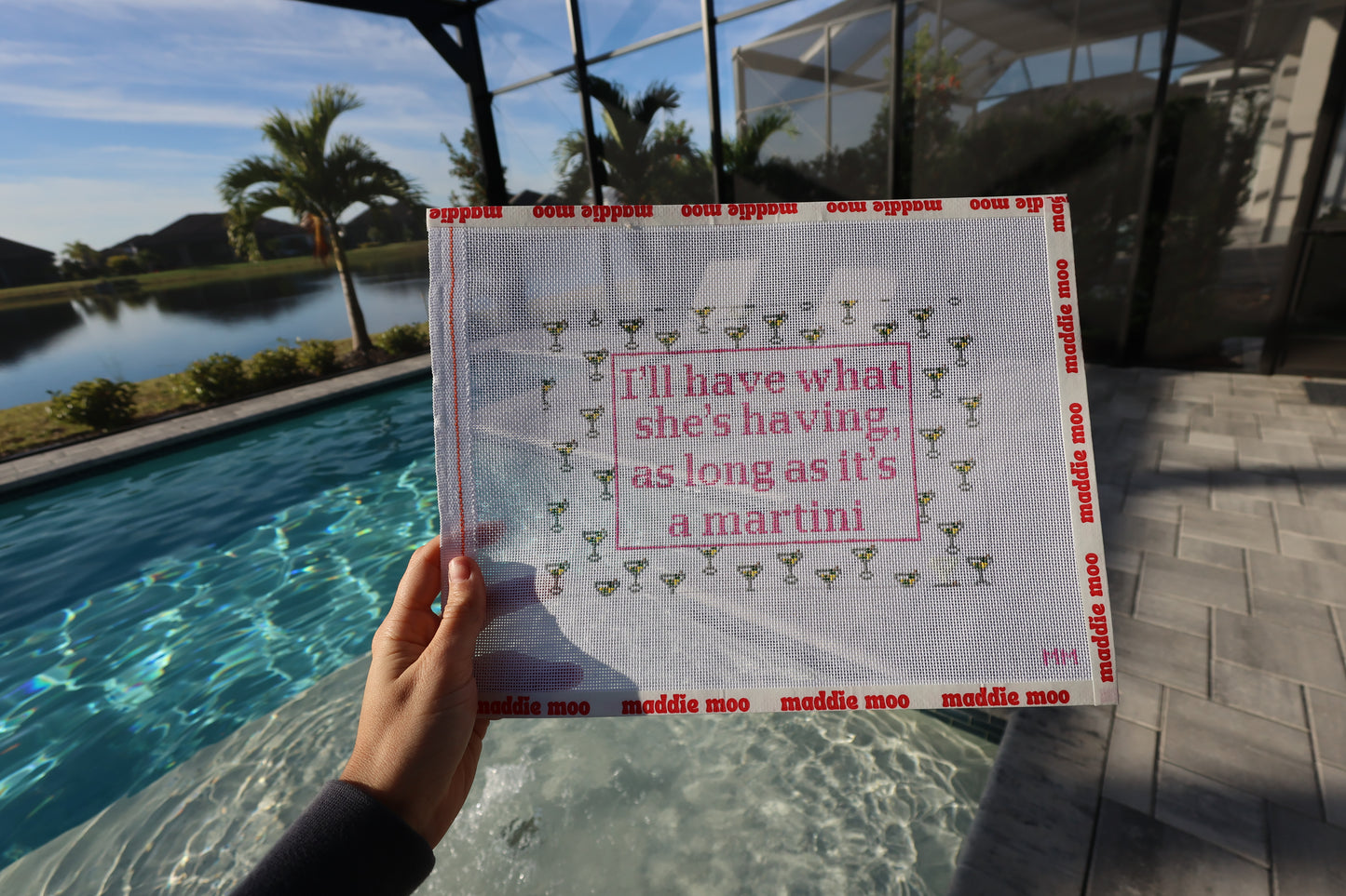 Hand-painted I'll Have What She's Having needlepoint canvas with a dirty martini illustration on 13 mesh, 12 by 8 inches, by Maddie Moo Needlepoint. Held by poolside.