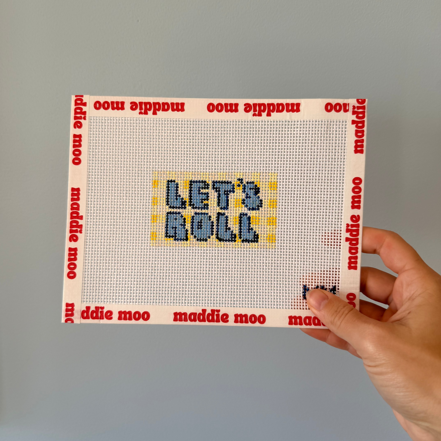Hand holding a cross-stitched panel with 'LET'S ROLL' text against a plain background